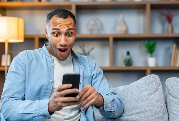 Man in casual wear sitting on sofa surprised by smartphone screen content indoors