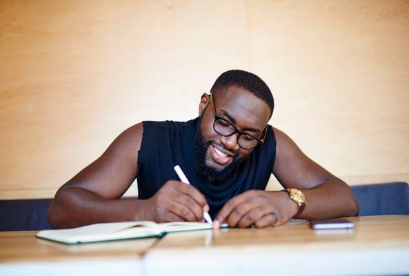 Smiling man wearing glasses writing in a notebook at a wooden desk indoors